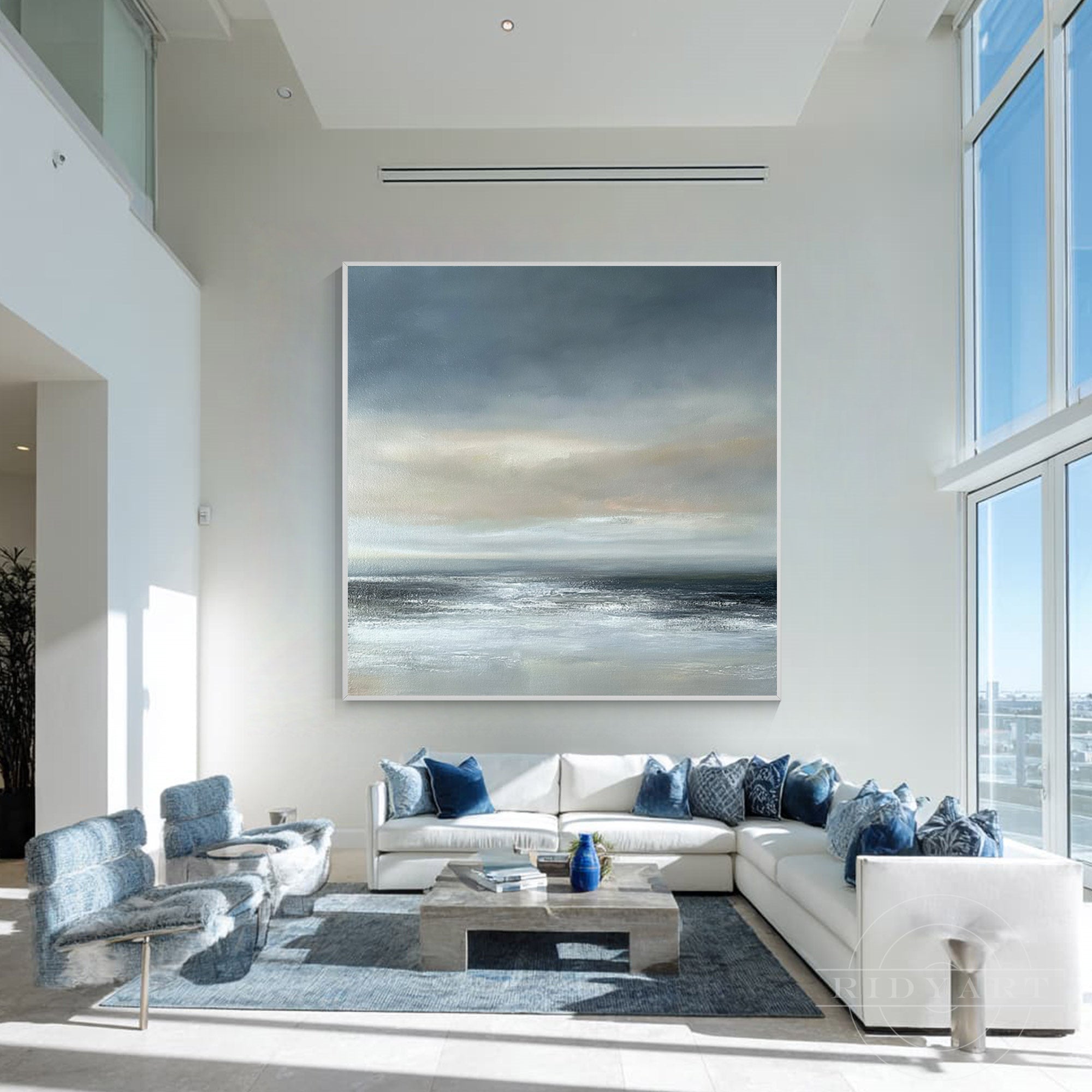 Extra large square abstract ocean canvas art framed in white, hanging high above a white sectional sofa and blue armchairs in a modern open-concept room.