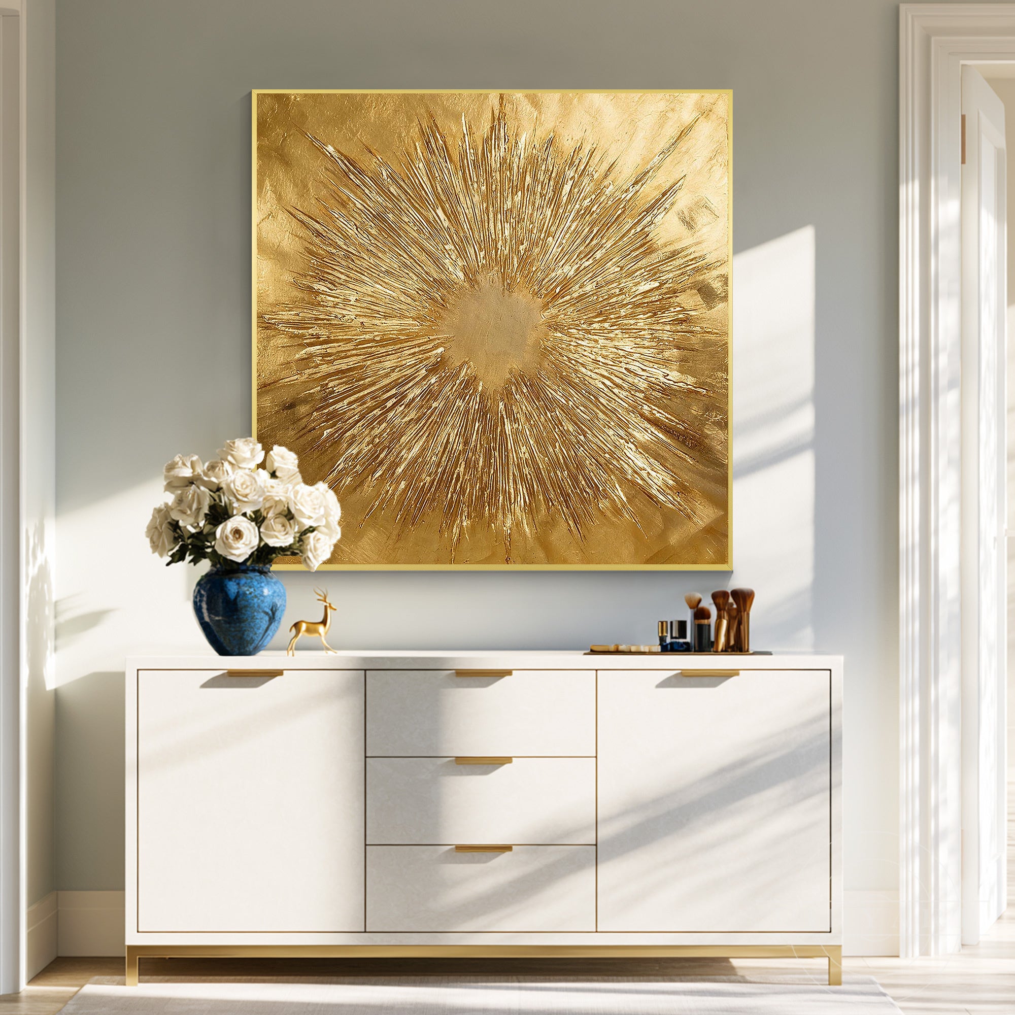 Square gold leaf sunburst painting in a gold frame hanging above a white sideboard with a blue vase of white flowers and makeup accessories.
