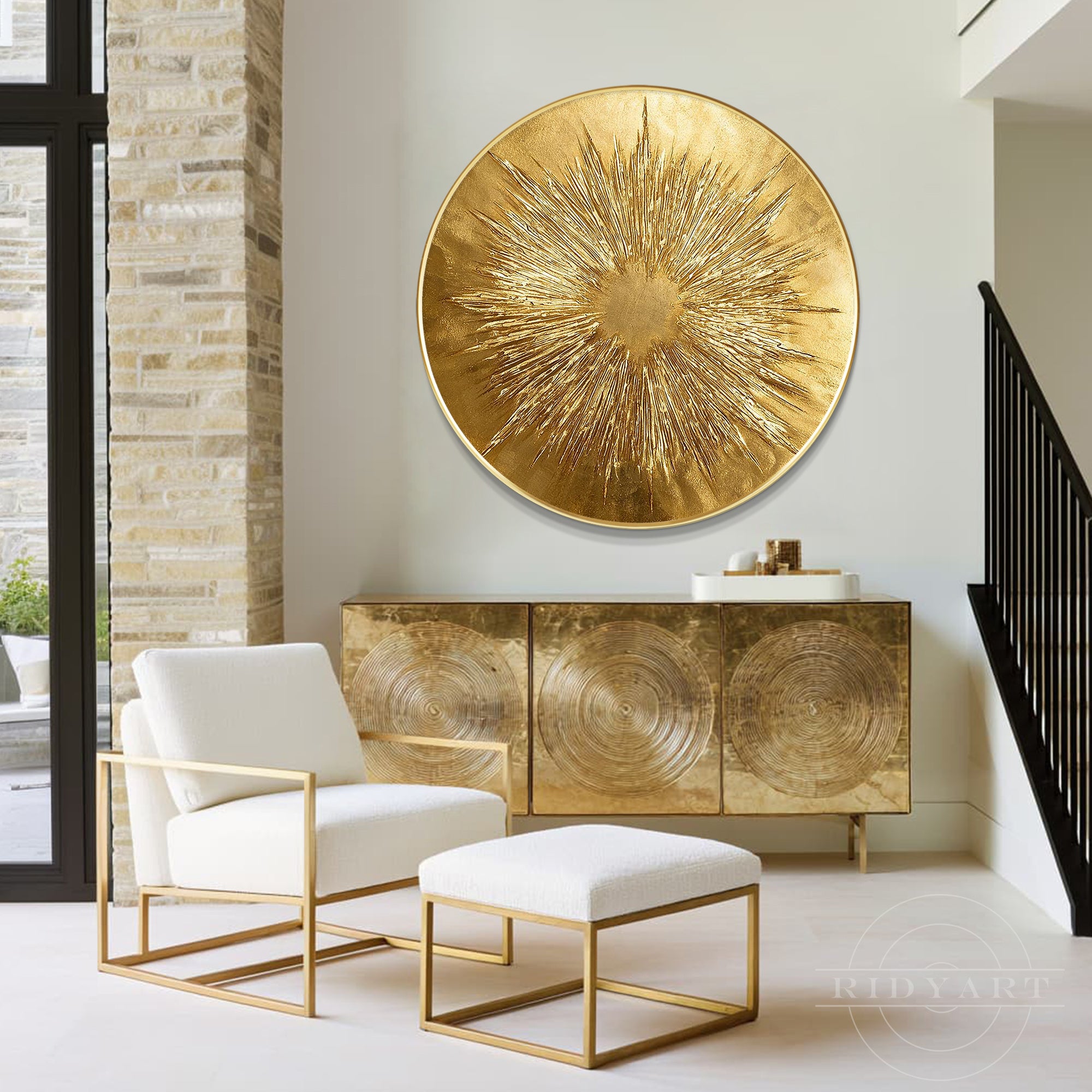 Circular gold textured starburst painting hanging above a metallic brass sideboard cabinet next to a white armchair and staircase.
