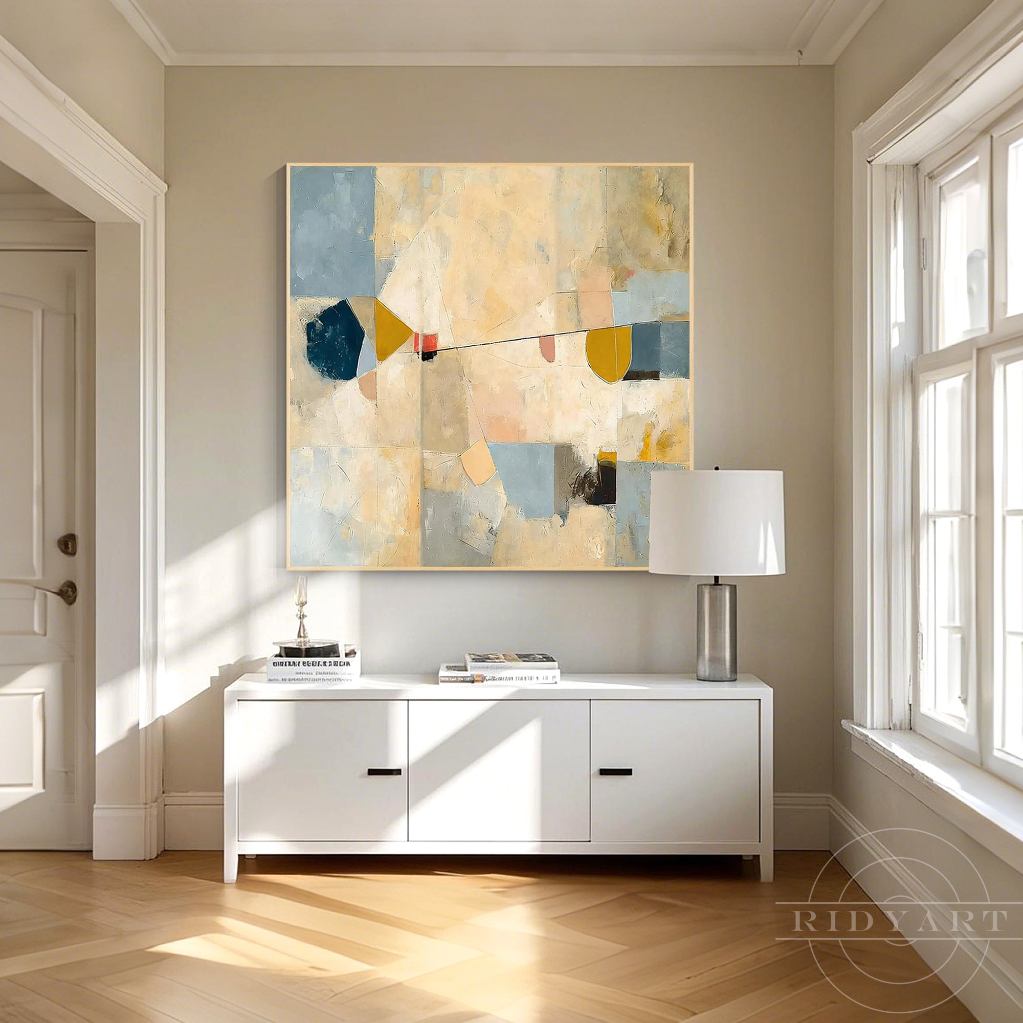 Framed mid-century warm beige abstract canvas artwork hanging above a white sideboard cabinet in a bright entryway hallway.