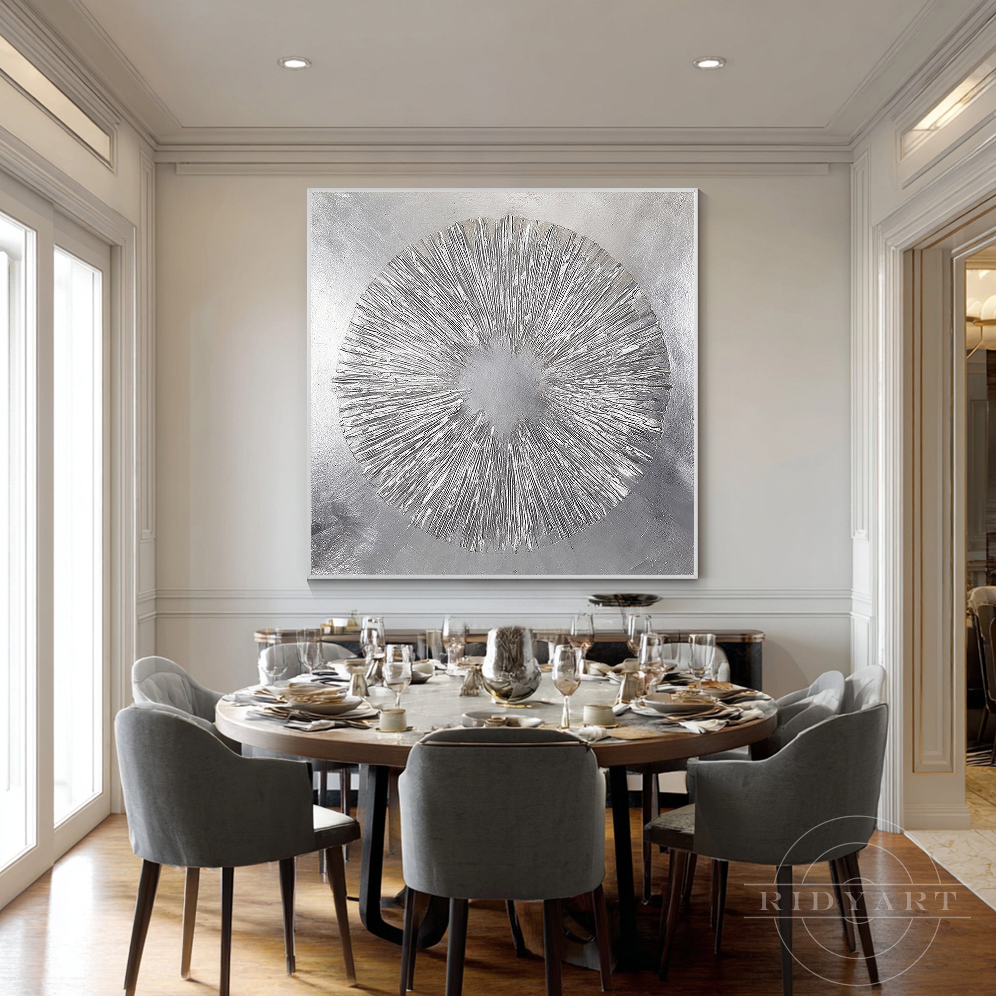 Square silver abstract burst art mounted on a dining room wall above a round dining table surrounded by grey velvet chairs.