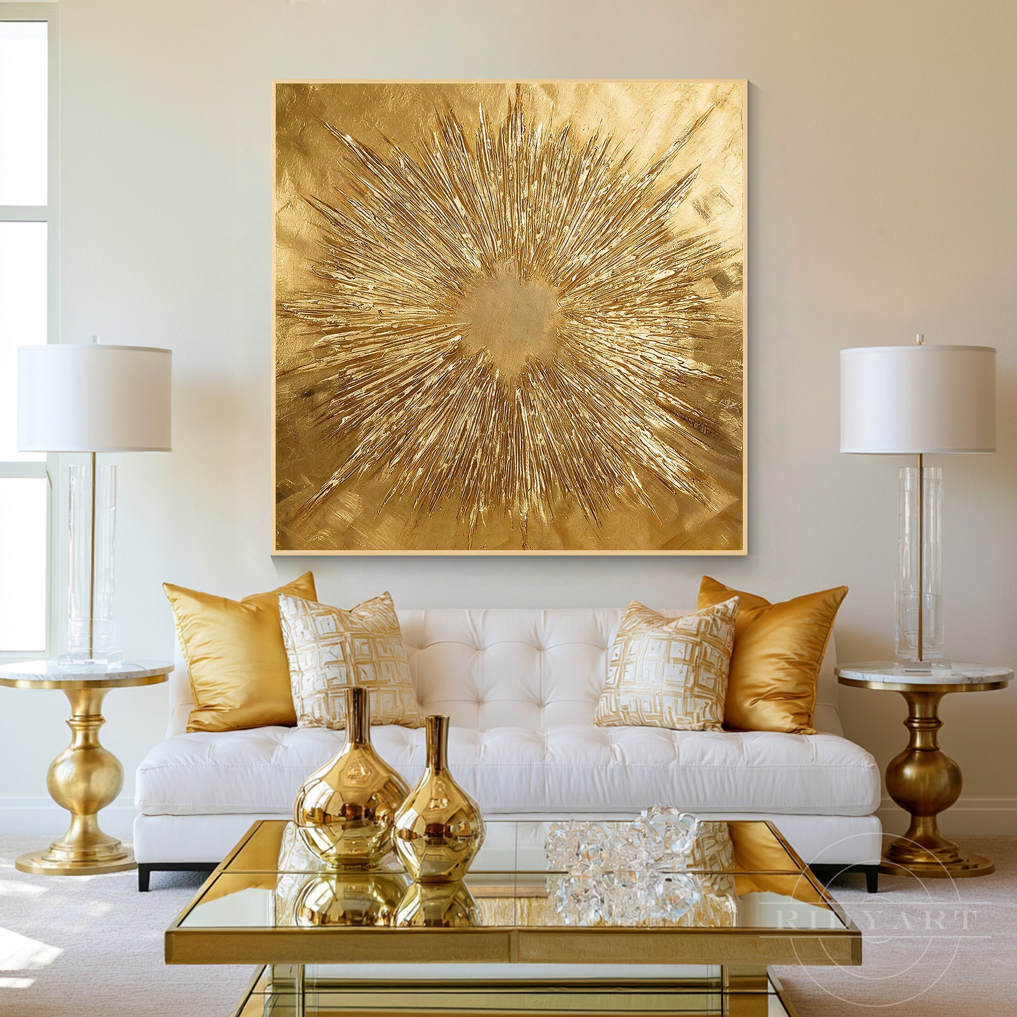 Square textured gold foil burst art displayed over a white tufted sofa with gold throw pillows and brass side lamps in a glamorous living room.