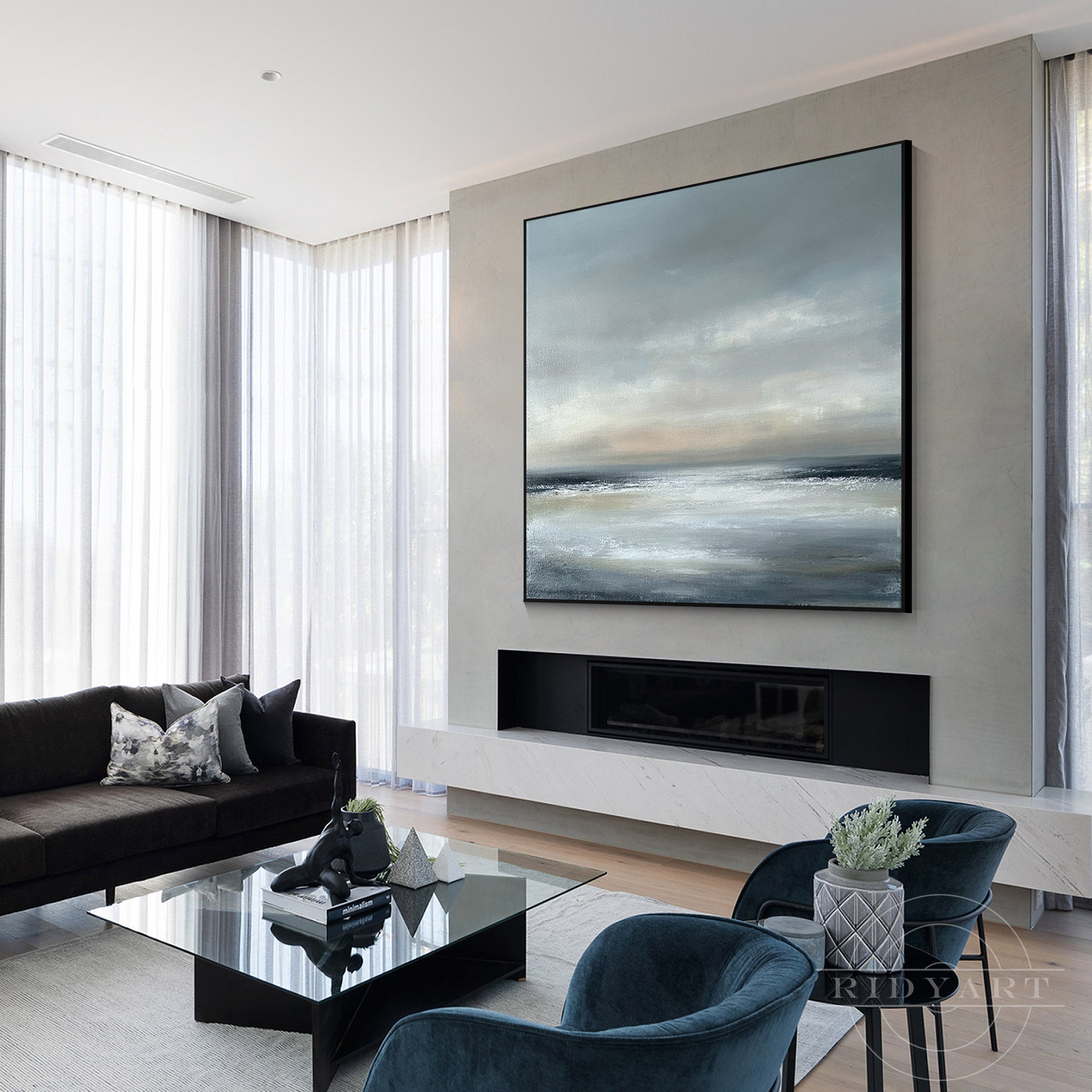 Square abstract ocean seascape canvas painting framed in black, displayed above a modern fireplace with a dark sectional sofa and blue velvet armchairs.
