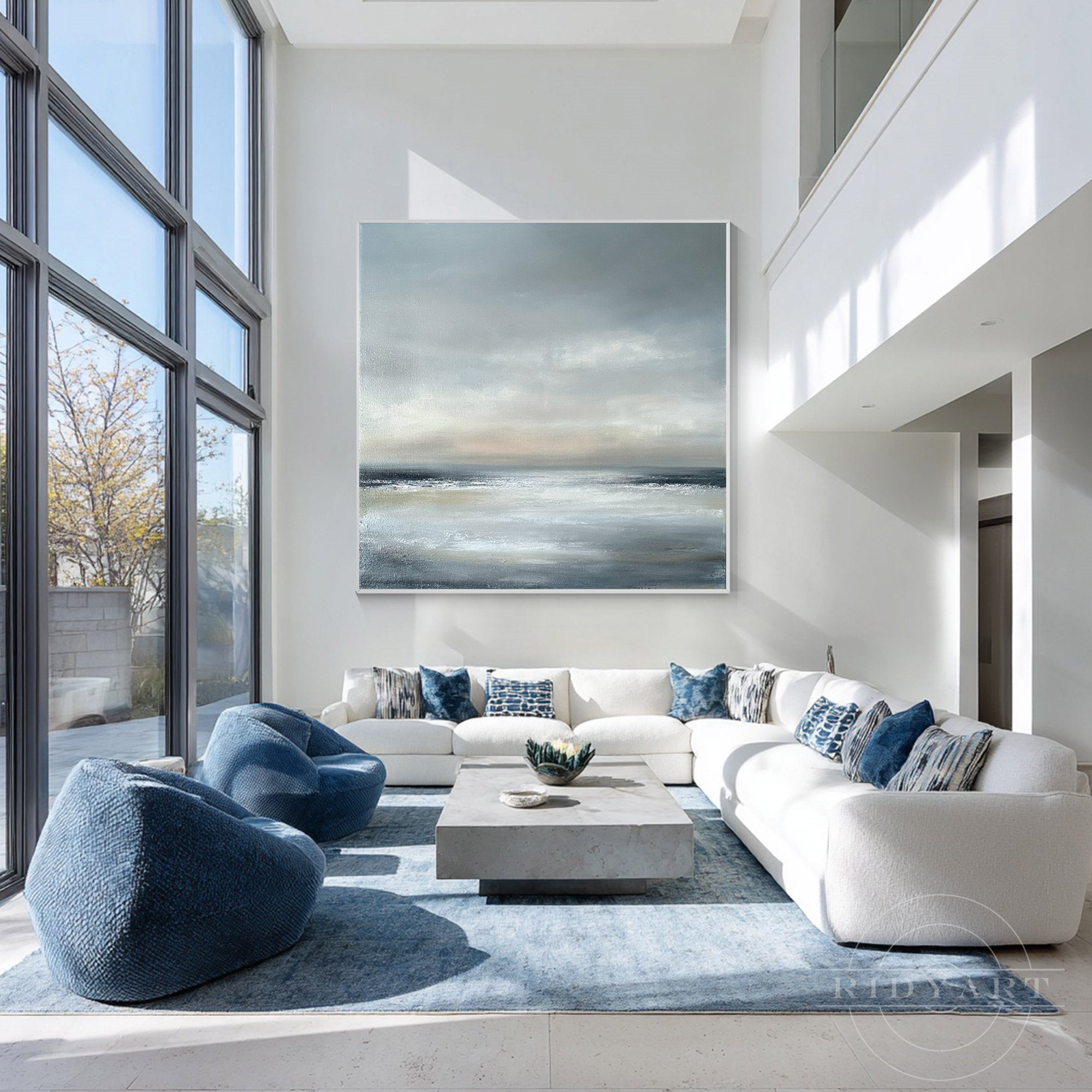 Square neutral grey and blue coastal horizon canvas painting mounted on a high-ceiling white wall above a large white sectional couch and blue beanbags.