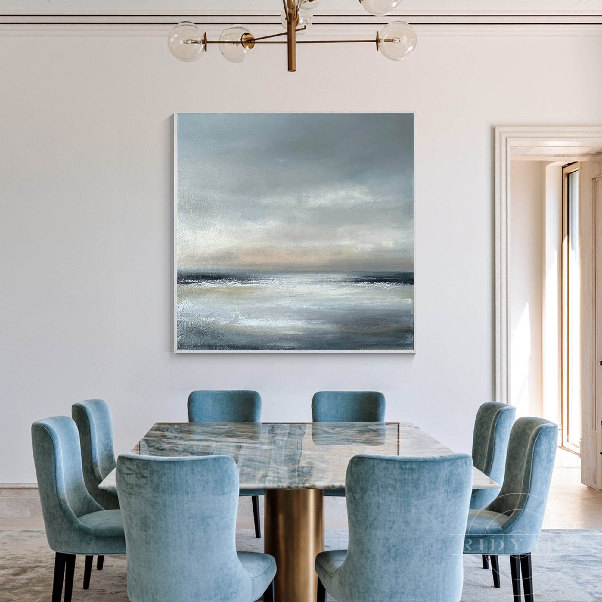 Square abstract grey and pale blue seascape art hanging in an elegant modern dining room above a marble table with blue velvet chairs.