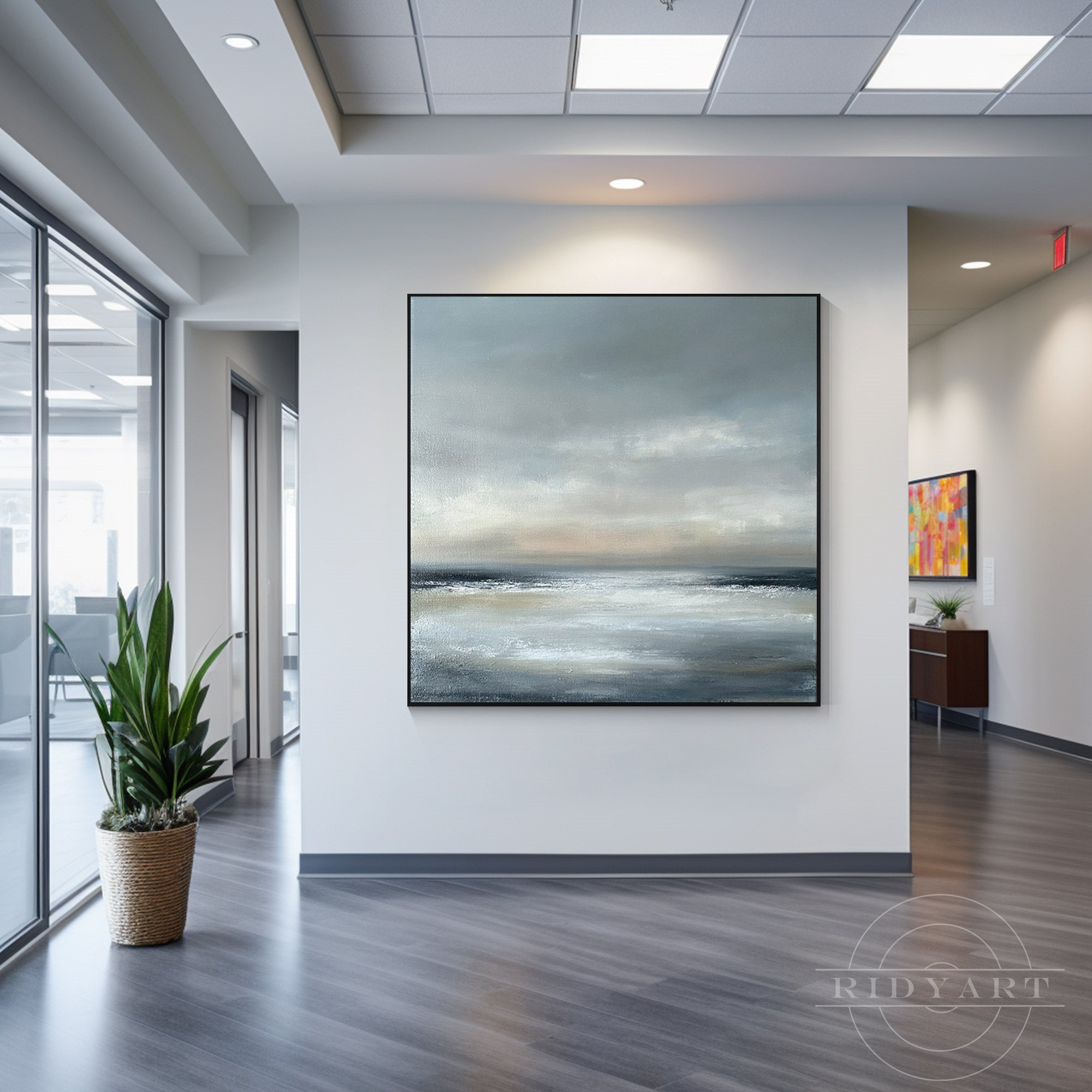 Square moody seascape canvas painting framed in black mounted on a white accent wall in a modern office hallway next to a potted floor plant.