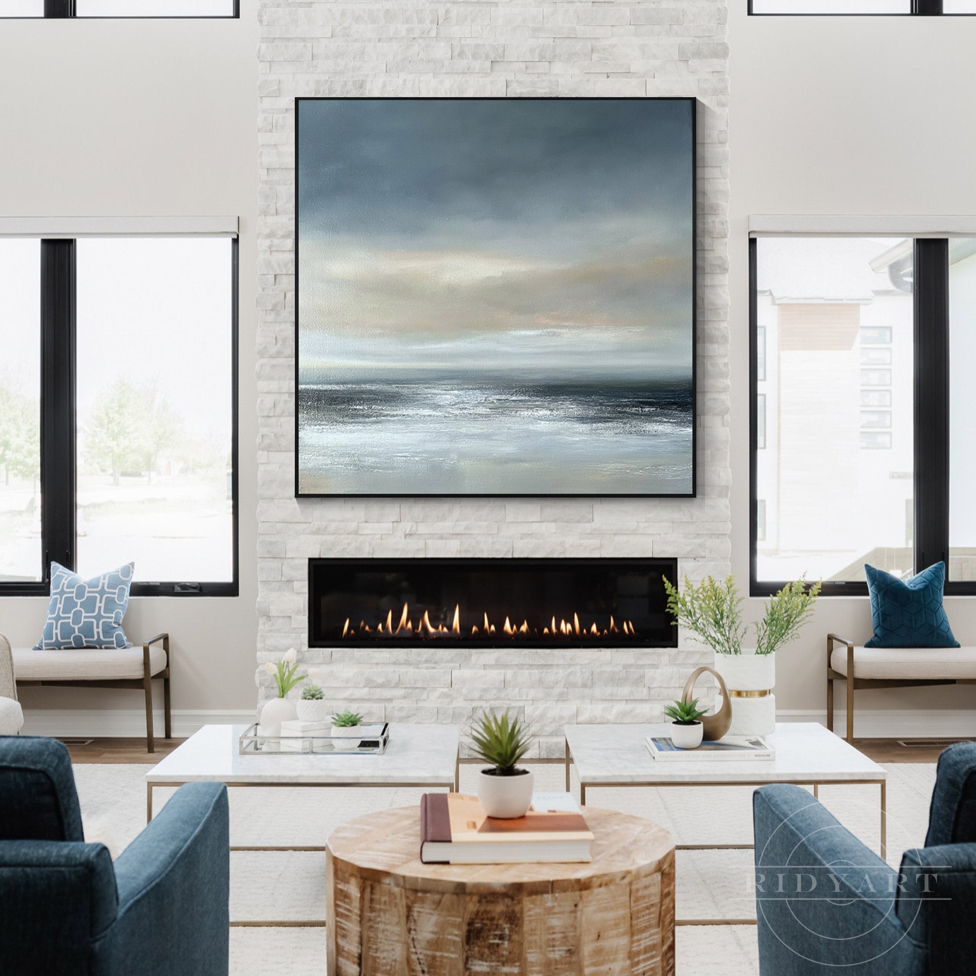 Large square ocean landscape painting centered over a modern white stone fireplace in a bright living room setup.